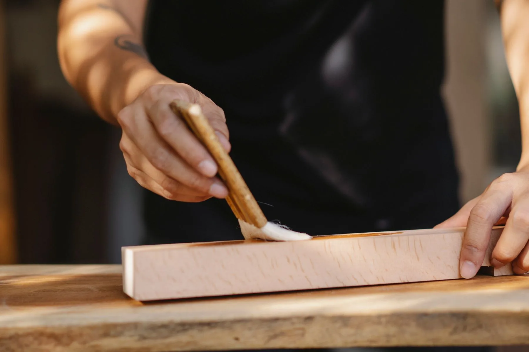 Manos de un artesano aplicando barniz con un pincel a una pieza de madera.
